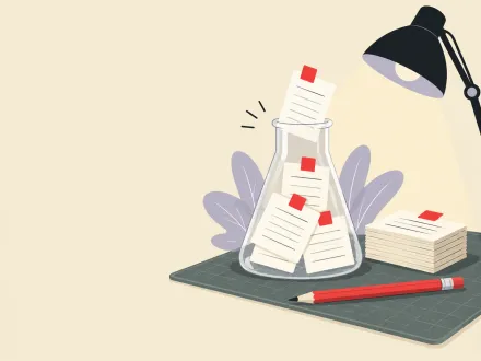 A glass lab flask holding index cards sits on a cutting mat beside a stack of cards, a red pencil and a desk lamp.